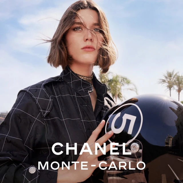 Portrait of a woman in a black jacket holding a racing helmet, with palm trees and blue sky in the background. Fashion or editorial shot.