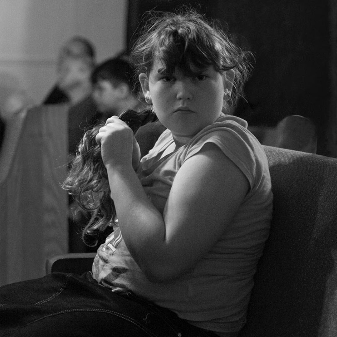 Black and white portrait of a child seated, looking at the camera, and holding their hair in a casual, indoor setting with background figures.