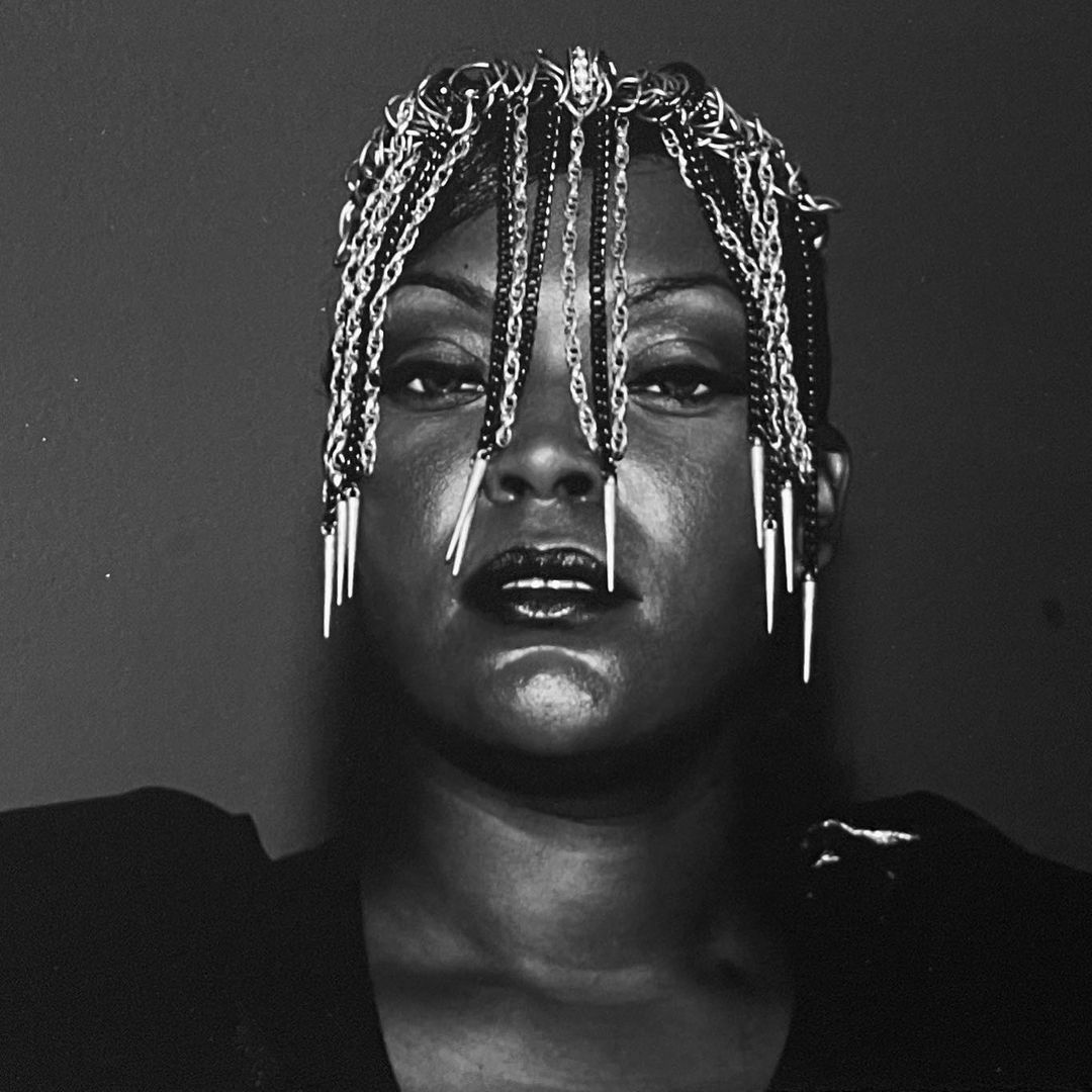 Close-up black and white portrait of an African-American woman wearing a metal fringe headpiece, looking at the camera against a dark background.