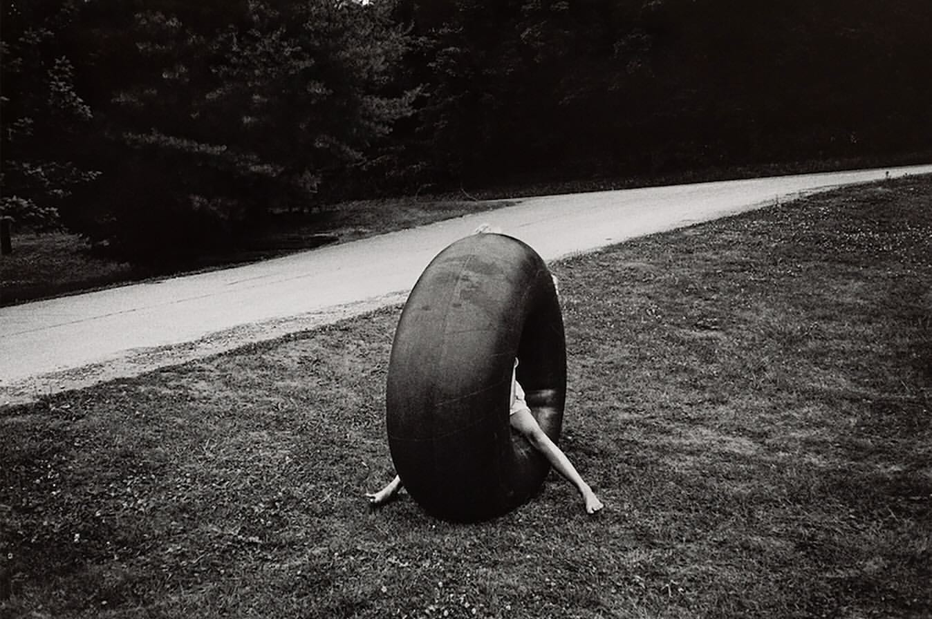 Monochrome image of a person inside a large tire, set on grassy terrain with trees and a road in the backdrop, creating a surreal scene.