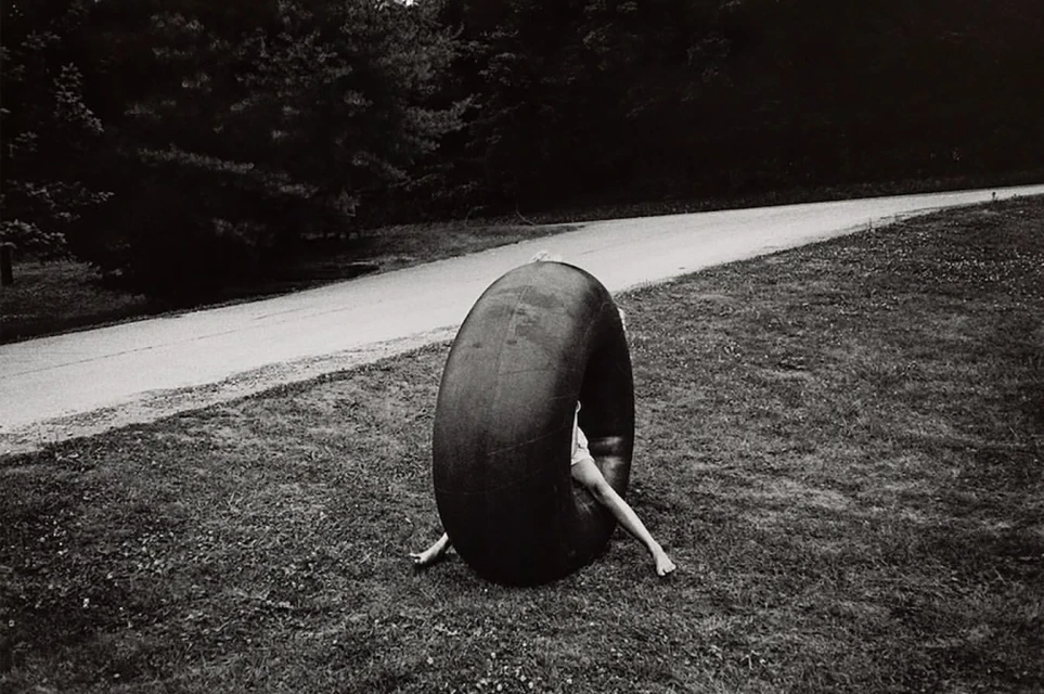 Monochrome image of a person inside a large tire, set on grassy terrain with trees and a road in the backdrop, creating a surreal scene.