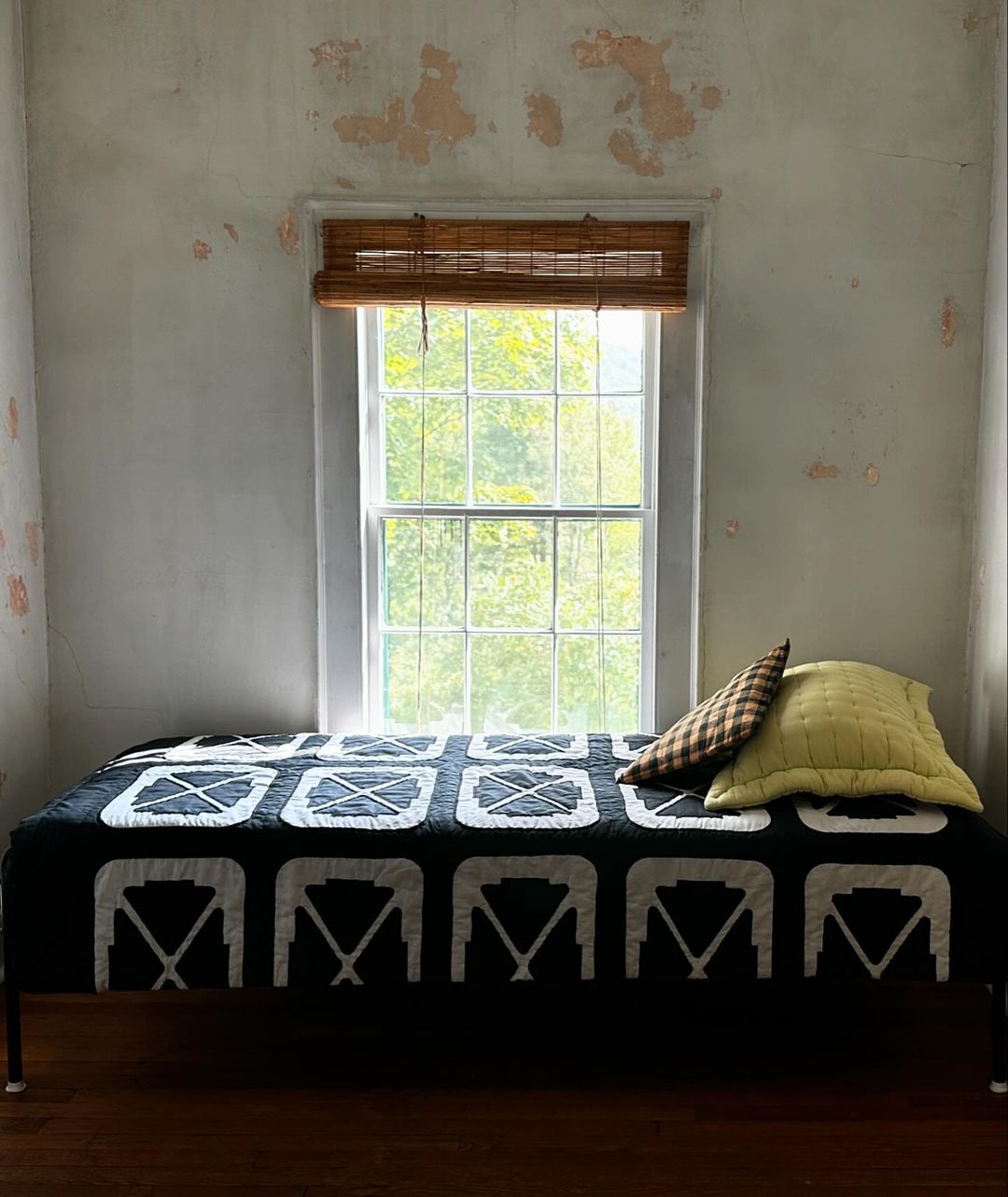 Interior view features a daybed with black and white patterned quilt, positioned beneath a window with a bamboo shade, next to pillows.