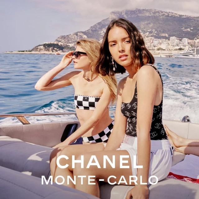 Two women wearing Chanel swimwear pose on a boat with a mountain and town in the background, capturing a luxury and fashionable aesthetic.