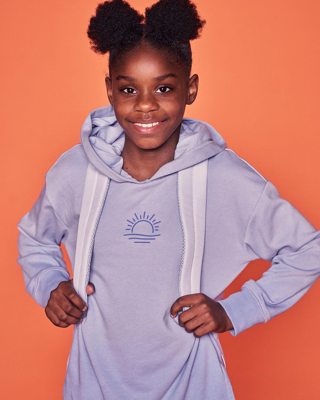 Young girl with afro puffs, smiling in a lavender hoodie dress against a bright orange studio backdrop.