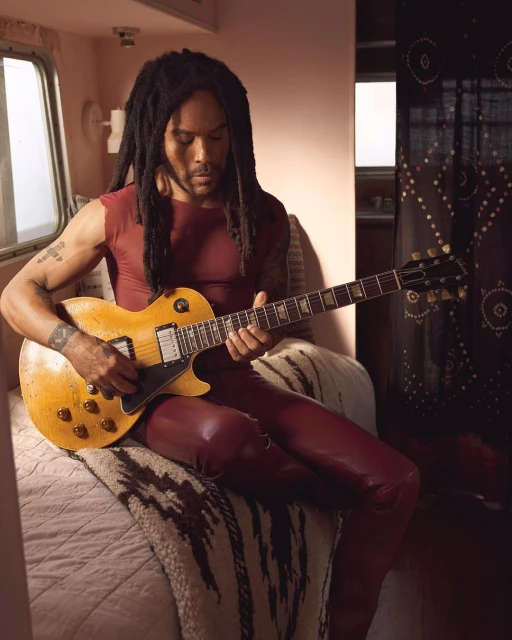 Adult male musician with dreadlocks, playing an electric guitar while sitting indoors on a bed. He is wearing a sleeveless top and leather pants.