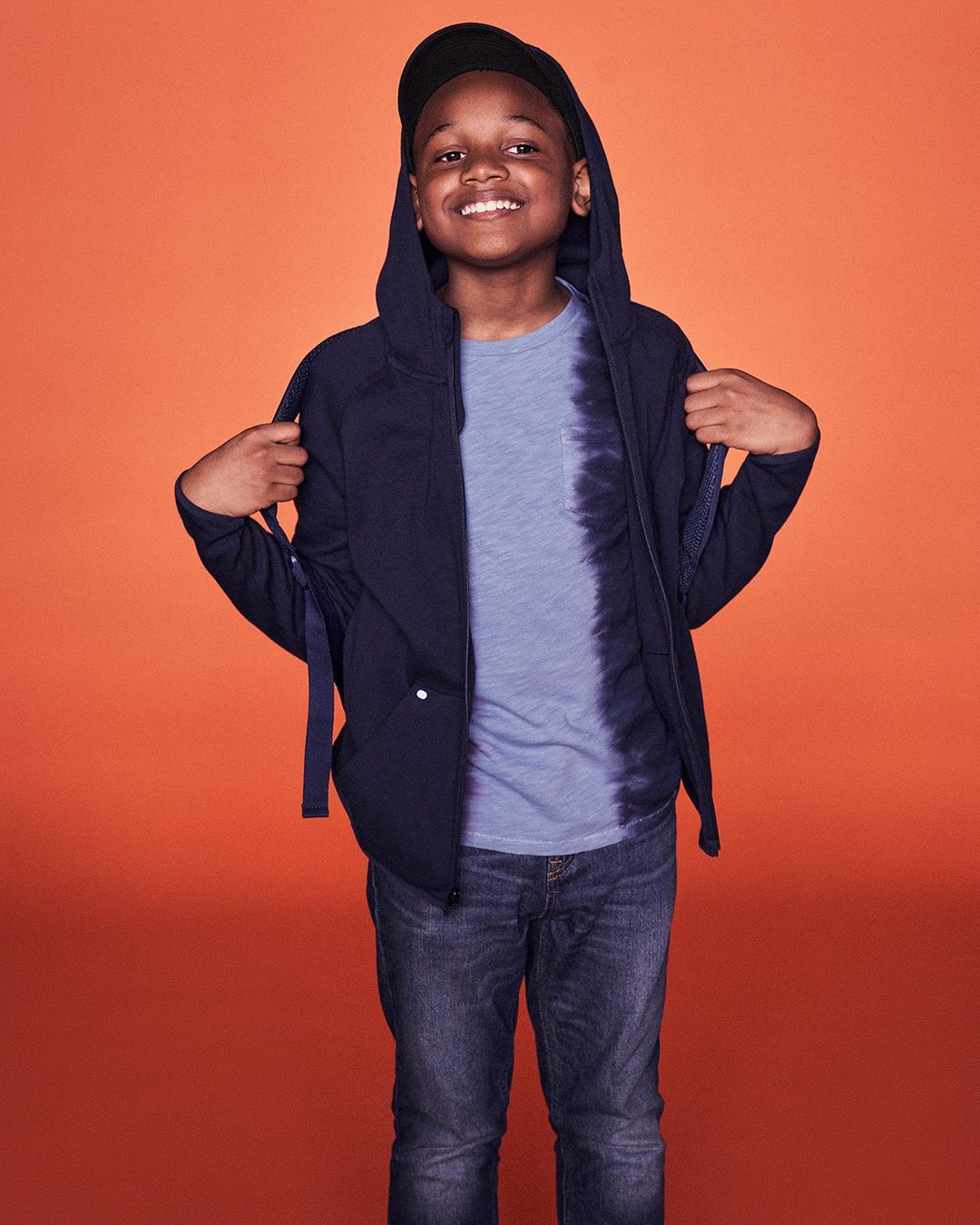 Young African-American boy stands smiling in a navy hooded zip-up jacket, light blue tie-dye shirt, jeans and baseball cap on orange backdrop.