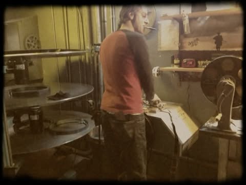 Man in red and black raglan works inside industrial setting near large metal discs and vintage fan with shelf and wall details.