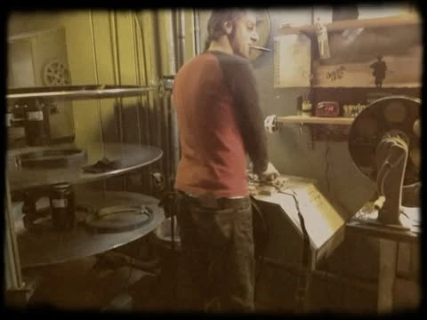 Man in red and black raglan works inside industrial setting near large metal discs and vintage fan with shelf and wall details.