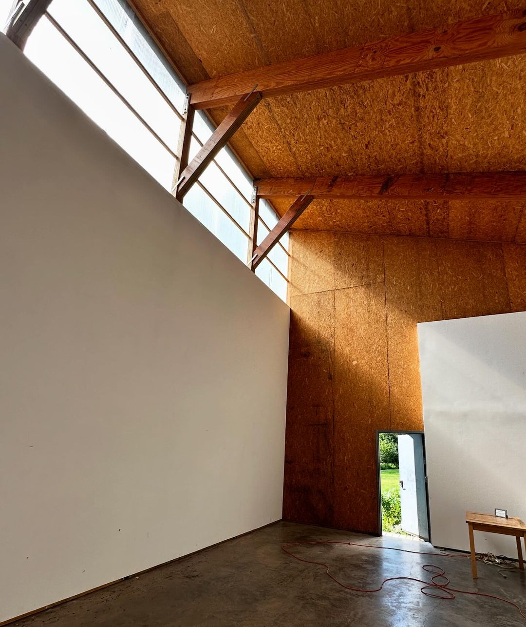 Spacious empty room with white walls and wood-paneled ceiling illuminated by natural light through large windows, a table, and open door.