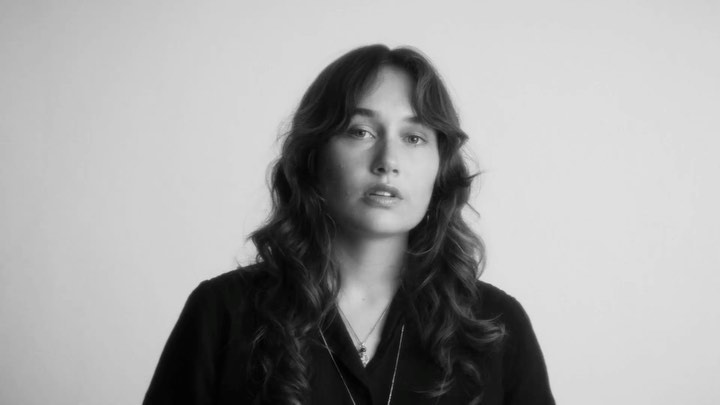 Monochrome portrait of a young adult woman with long curly hair, wearing a dark top and necklaces, against a plain white background.