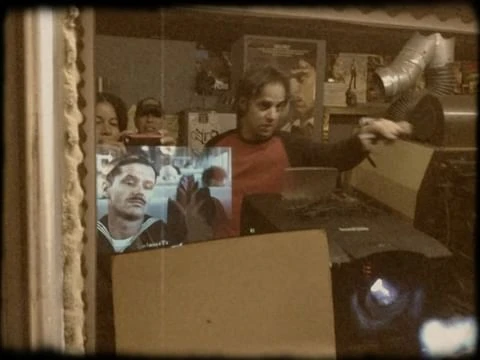 Indoor shot of two adult males, an adult female, and a child. One man is opening a box. A TV screen shows a sailor's image.