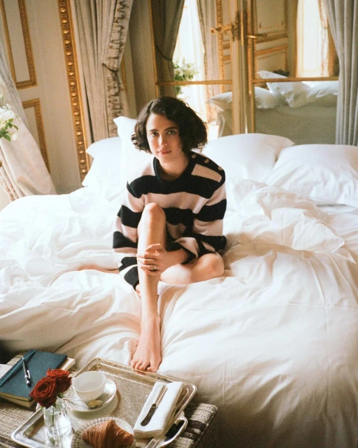 Woman in striped shirt sits on a white bed near a breakfast tray with roses, croissant, and coffee in a bright interior.