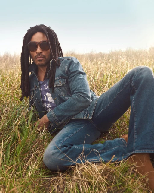 Adult male with dreadlocks and sunglasses lies in a grassy field wearing a denim jacket, t-shirt, and jeans in an outdoor shot.