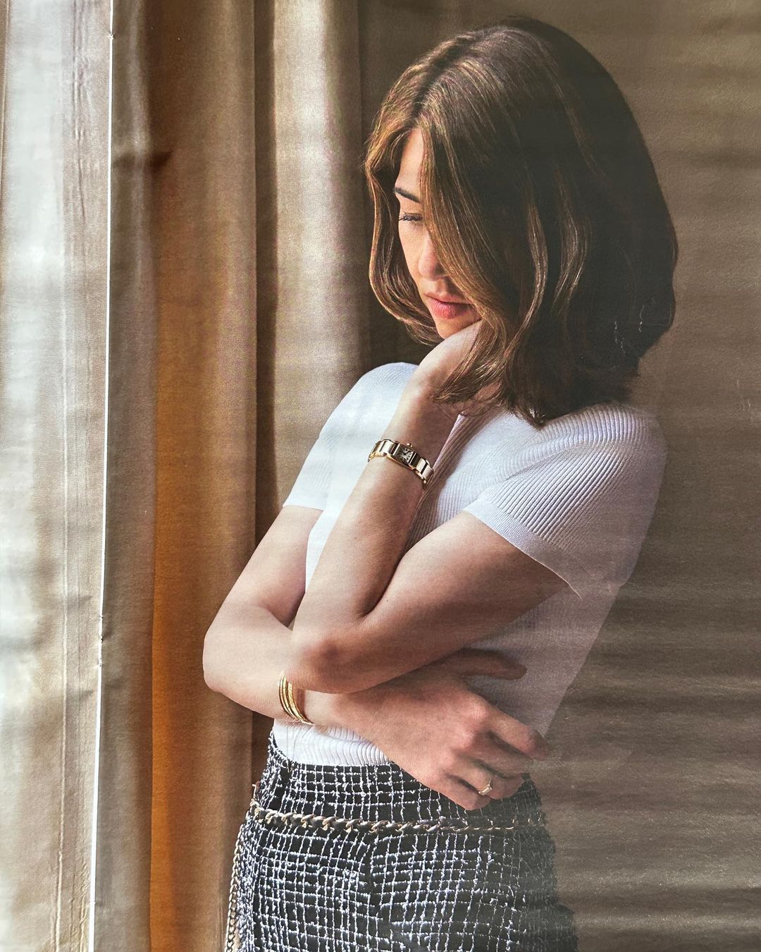 Woman in white top and patterned skirt, looking down with crossed arms, indoors near window with curtains.