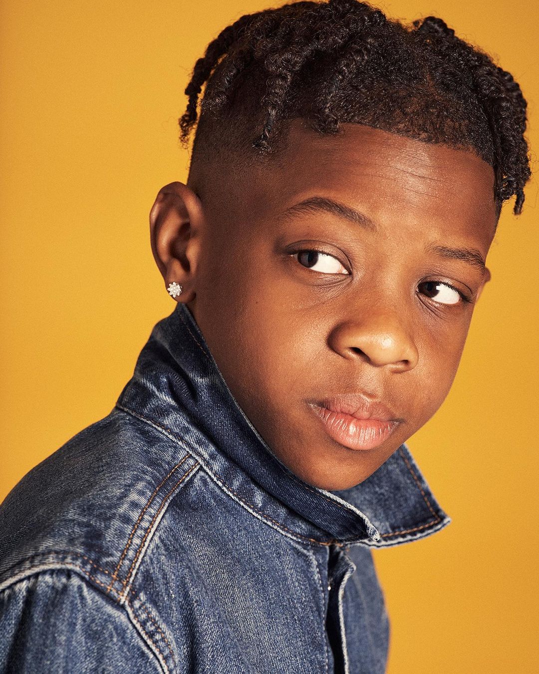 Portrait of a serious young African-American boy in a blue denim jacket looking to the side against a yellow background in a studio shot.