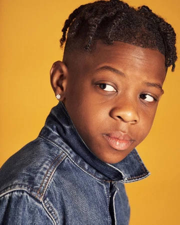 Portrait of a serious young African-American boy in a blue denim jacket looking to the side against a yellow background in a studio shot.
