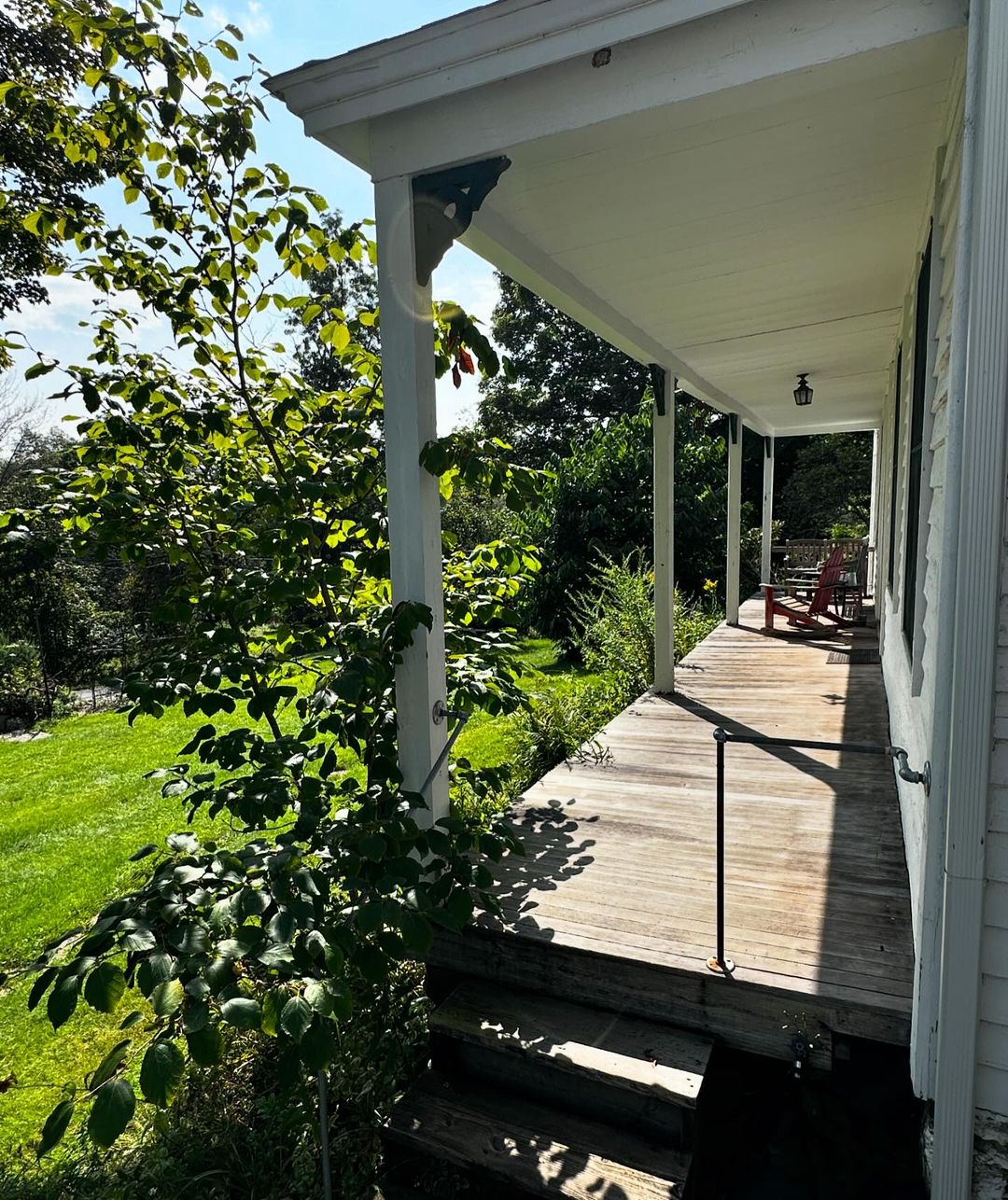 Outdoor porch scene with green lawn, foliage, white columns and trim, wooden decking, steps, black railing, and distant red Adirondack chairs.