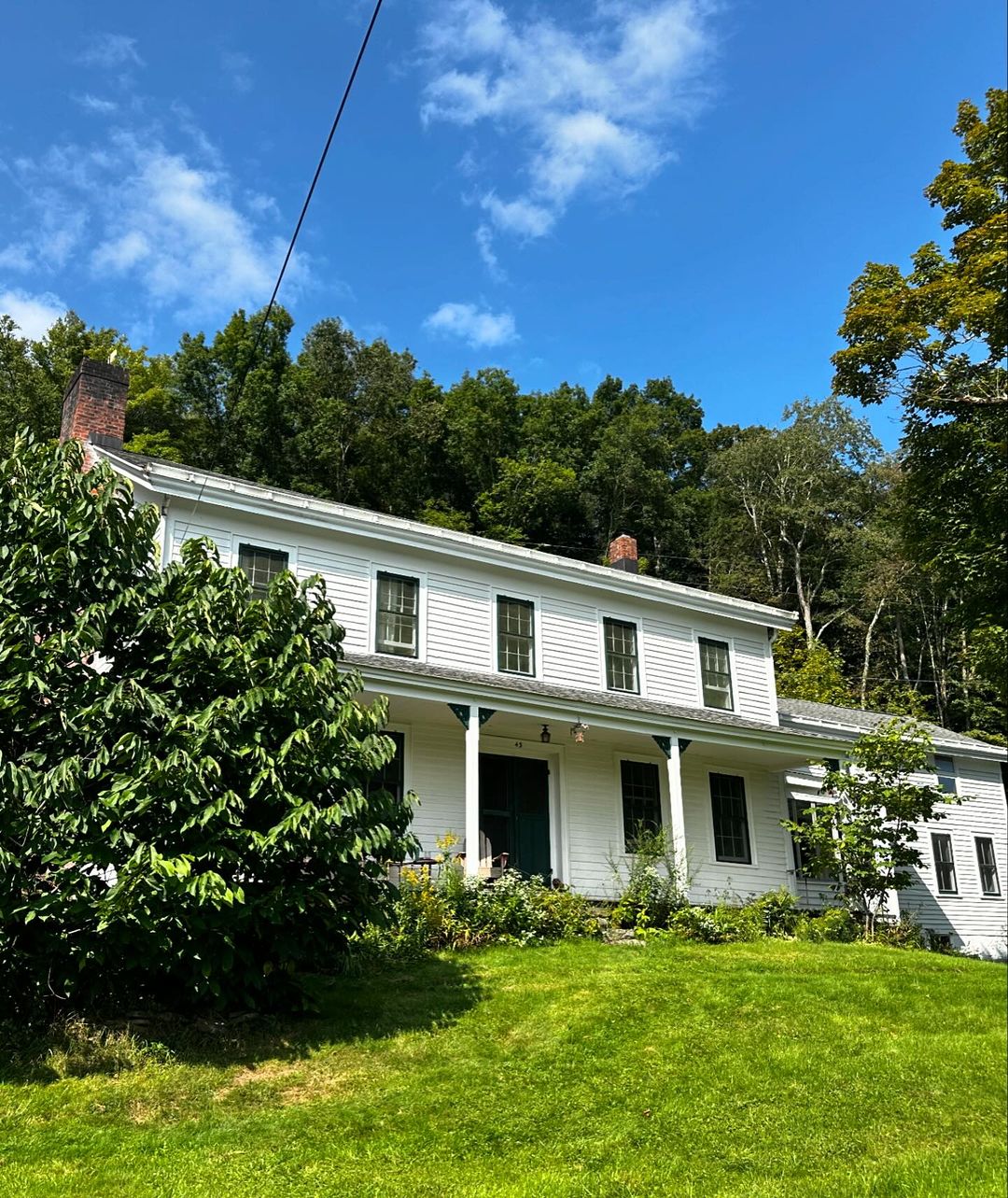 Two-story white farmhouse with a brick chimney under a bright blue sky with puffy white clouds surrounded by green trees and grass.