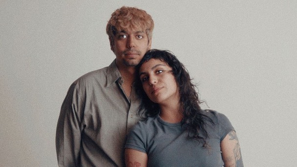 Portrait of a couple with warm skin tones in casual clothing. Woman leans her head on the man's shoulder against a plain background.