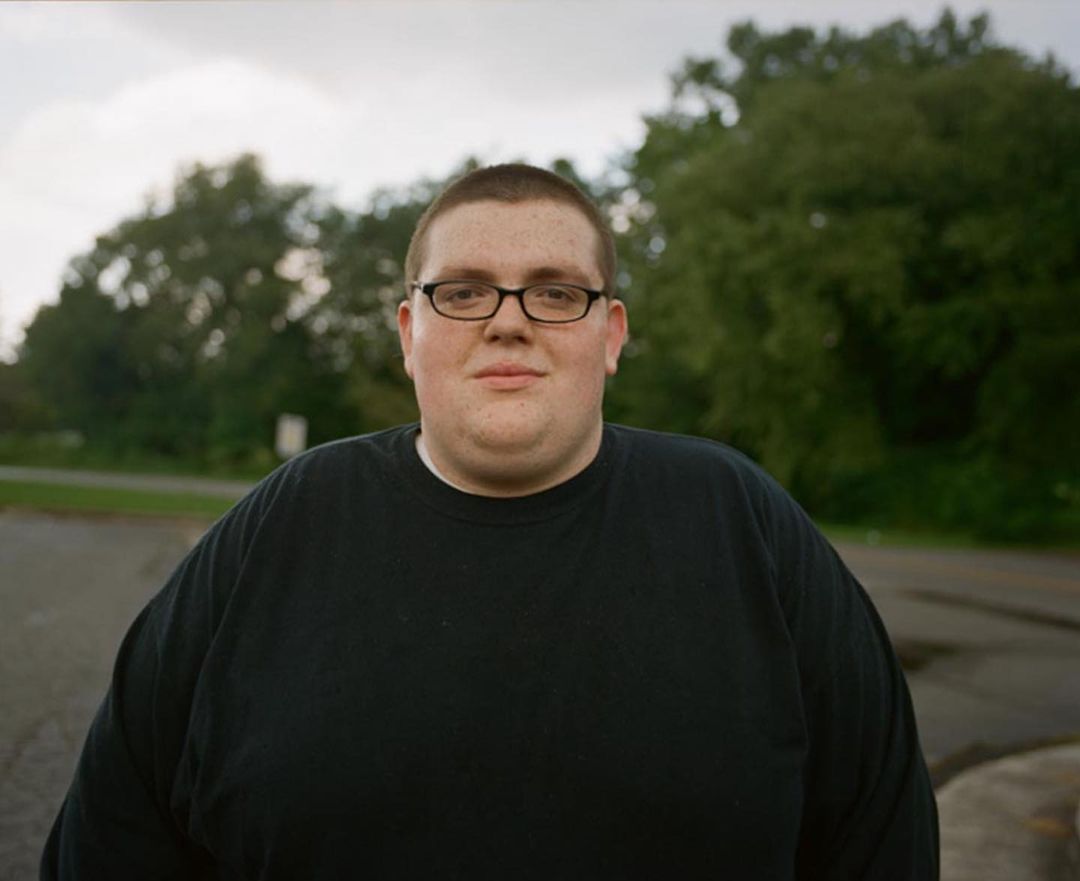Portrait of a young adult male with glasses and a black shirt outdoors against a blurred background of trees and road.
