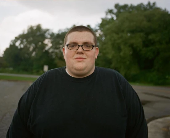 Portrait of a young adult male with glasses and a black shirt outdoors against a blurred background of trees and road.