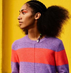 Profile portrait of a person with braided dark hair wearing a striped purple sweater against a solid yellow wall.