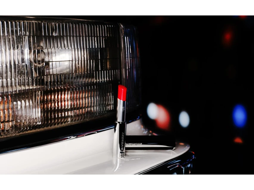 Red lipstick by Givenchy displayed on a white car with a headlight, set against a blurred background of red and blue bokeh lights.