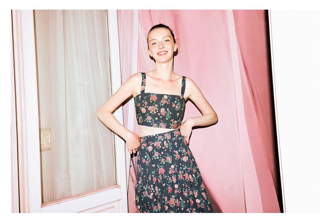 Smiling woman poses in a two-piece floral print outfit in front of a pink background with a neutral-toned door and window frame.