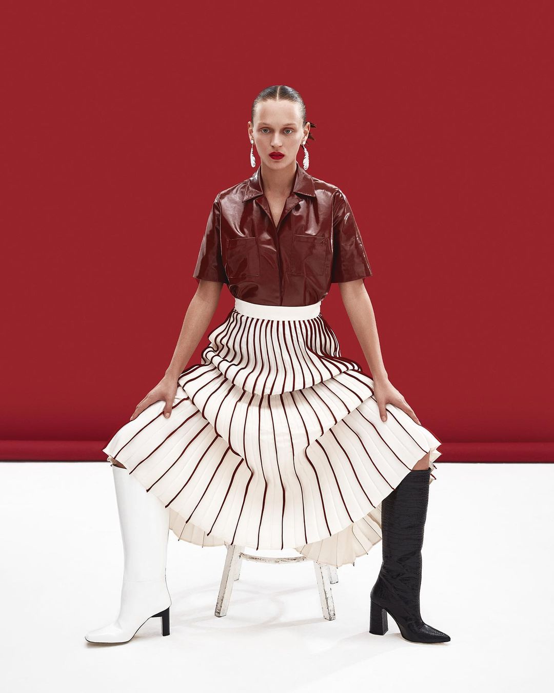 A woman with red lipstick and long earrings poses on a chair wearing a brown top, white skirt, and mismatched white and black boots.