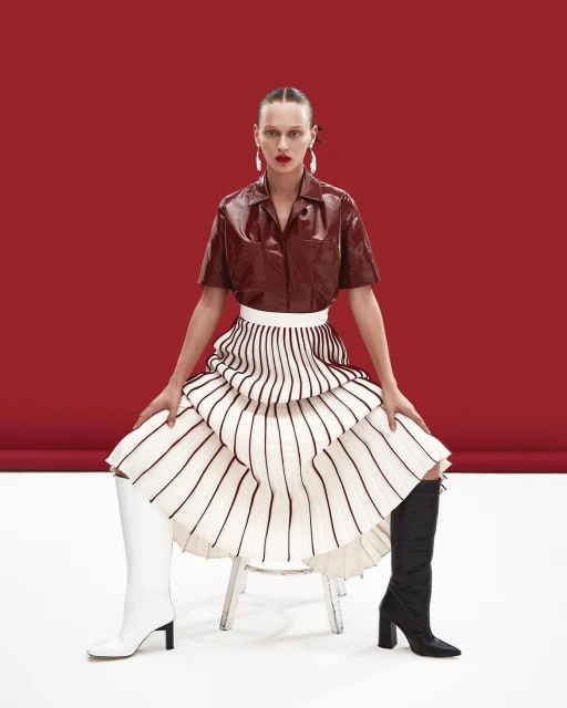 A woman with red lipstick and long earrings poses on a chair wearing a brown top, white skirt, and mismatched white and black boots.