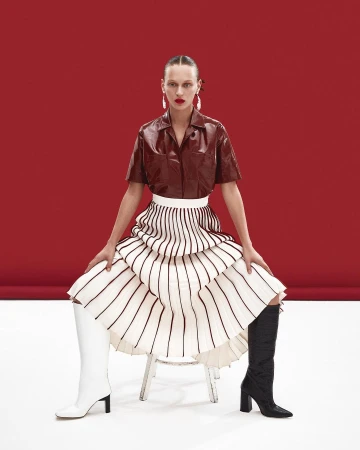 A woman with red lipstick and long earrings poses on a chair wearing a brown top, white skirt, and mismatched white and black boots.