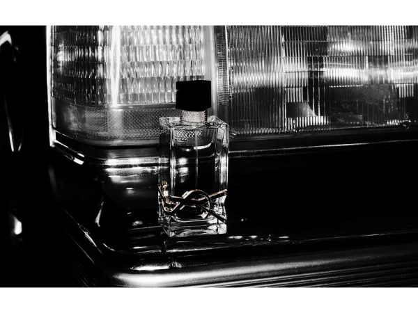 Elegant still life with a clear glass perfume bottle featuring a YSL logo, set against a black car grill and headlamp backdrop.