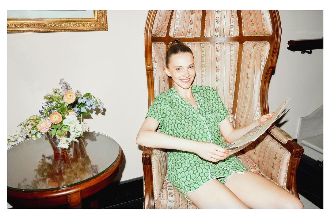 Woman in green patterned pajamas sitting in an ornate chair, reading a newspaper next to a table with a flower arrangement.