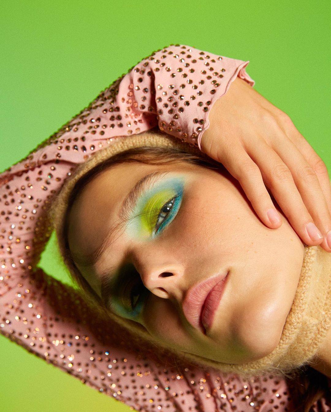 Close-up of a woman with vibrant blue and green eye makeup, wearing a light pink top with gold beads against a gradient green background.