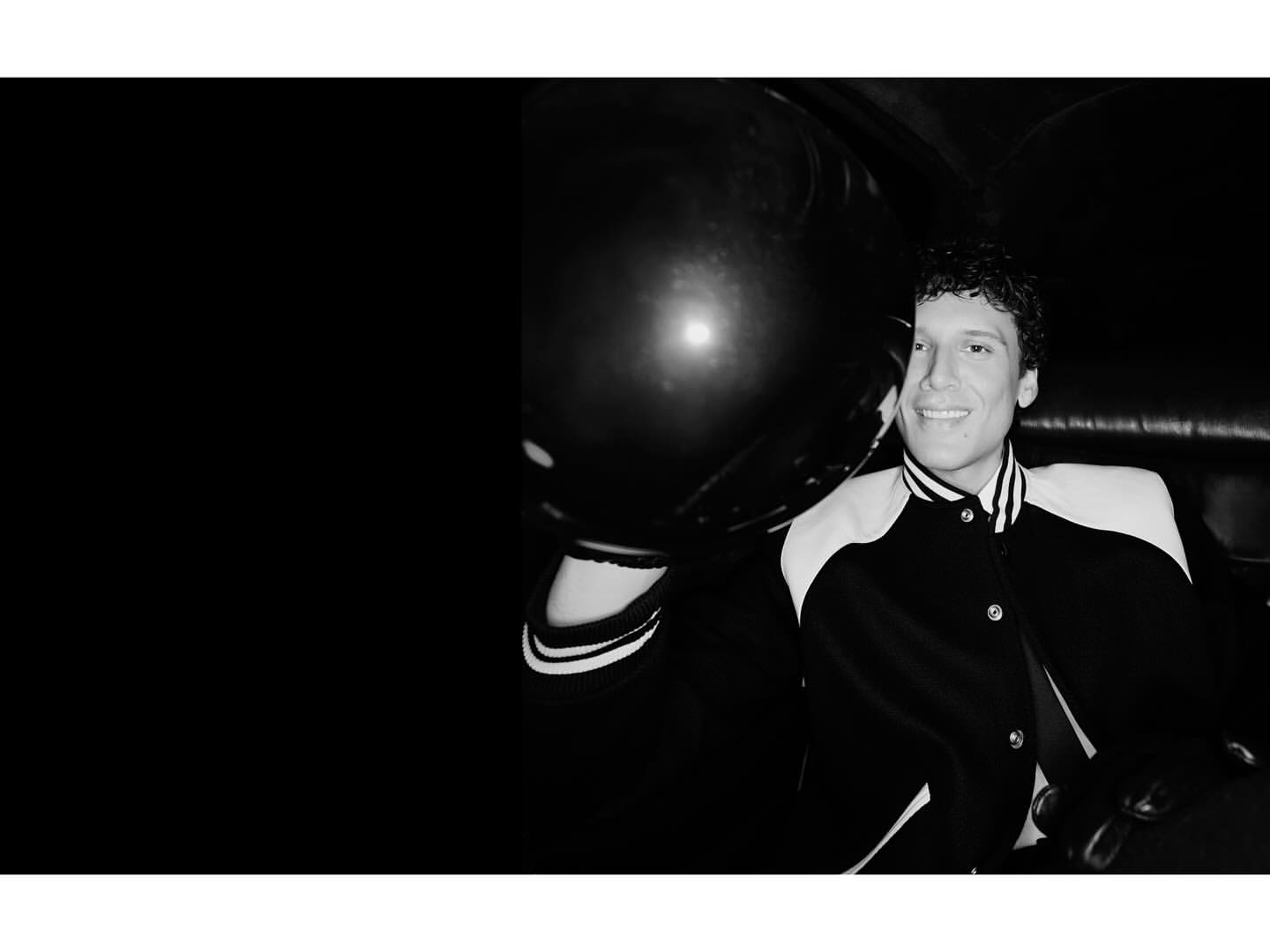 Portrait of a smiling adult male wearing a letterman-style jacket against a dark background with a large black balloon.
