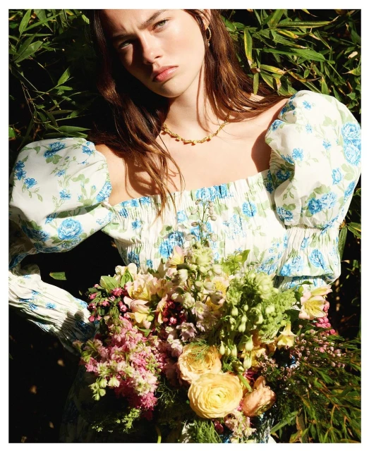 A woman in a floral top poses with a bouquet outdoors, bathed in natural light with a backdrop of green foliage, and a shallow depth of field.