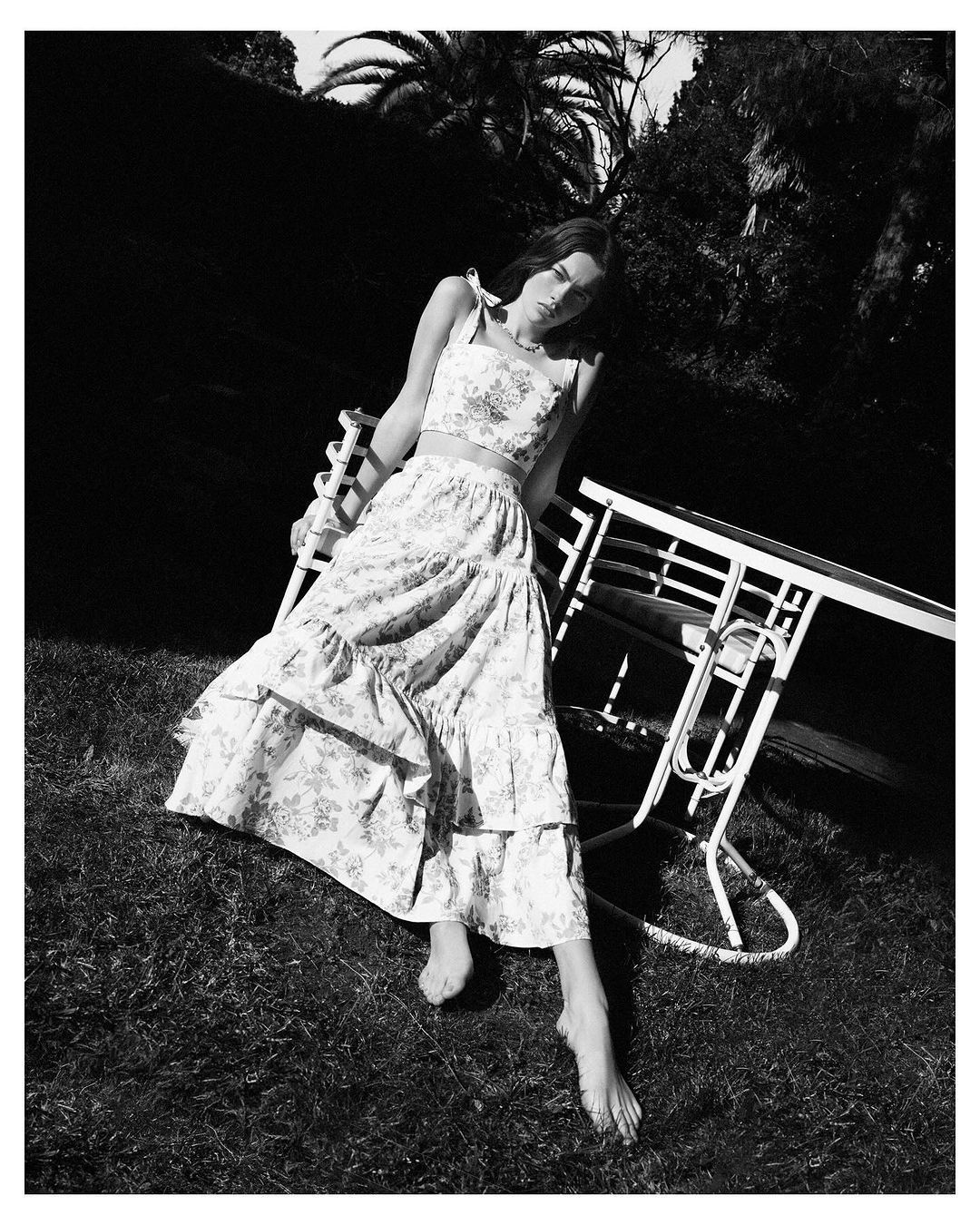 Young woman in floral print two-piece outfit poses outdoors sitting on white iron chair on grass in a monochrome photograph.