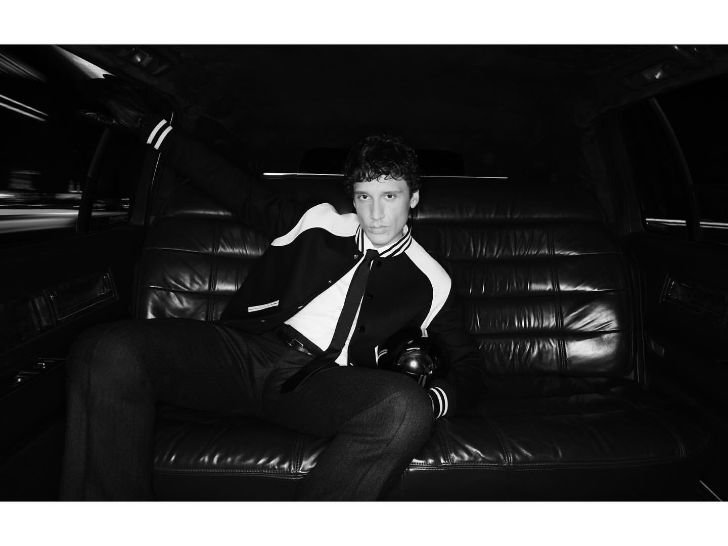 Monochrome portrait of a curly-haired young adult in a letterman jacket, seated in a leather car seat with dark interior.