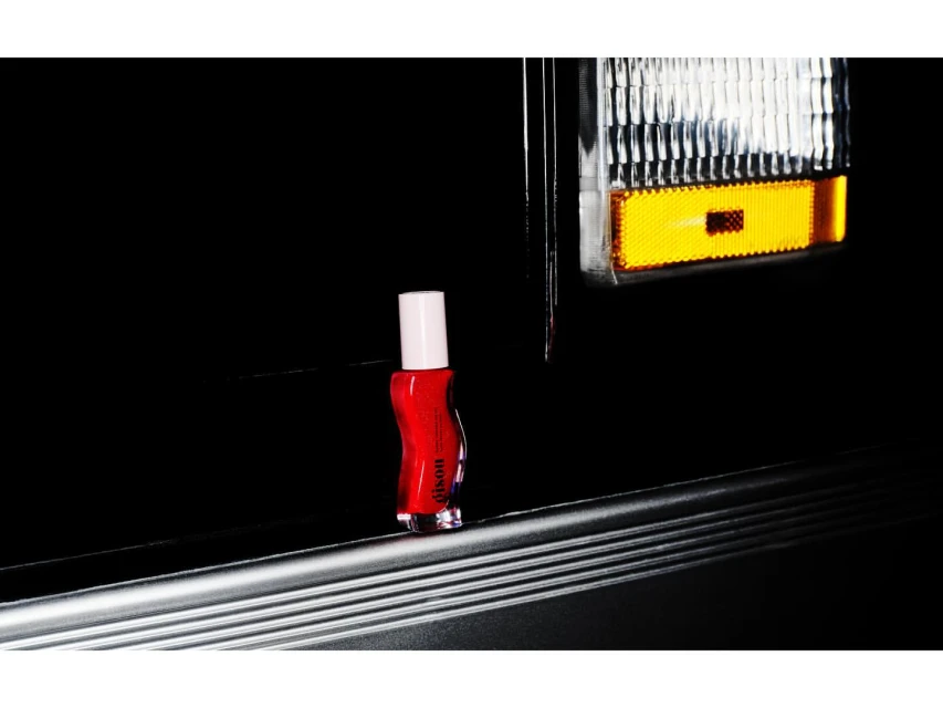 Product shot of red nail polish on a gray surface, dark background with a car headlight reflecting in the scene.