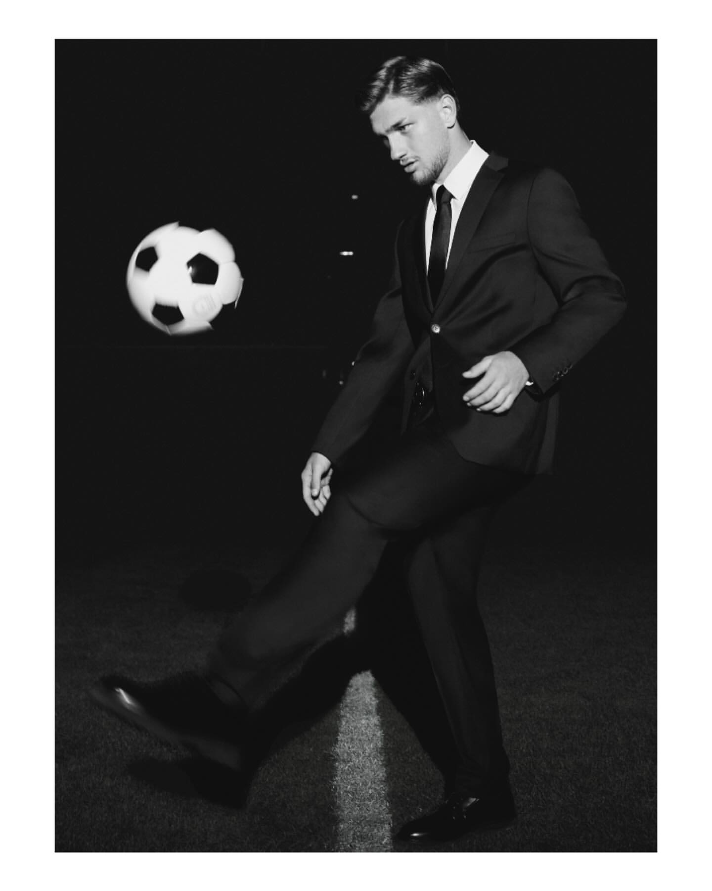 Monochrome image of a man in a suit kicking a soccer ball on a field, captured in a formal, low-angle shot against a black backdrop.