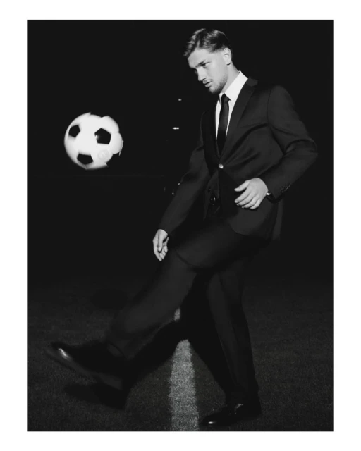 Monochrome image of a man in a suit kicking a soccer ball on a field, captured in a formal, low-angle shot against a black backdrop.