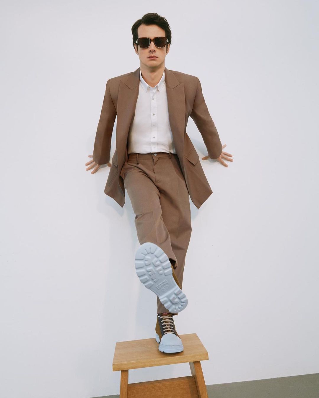 Stylish man in a brown suit posing on a wooden stool, showcasing a boot sole against a white backdrop in a fashion forward studio.