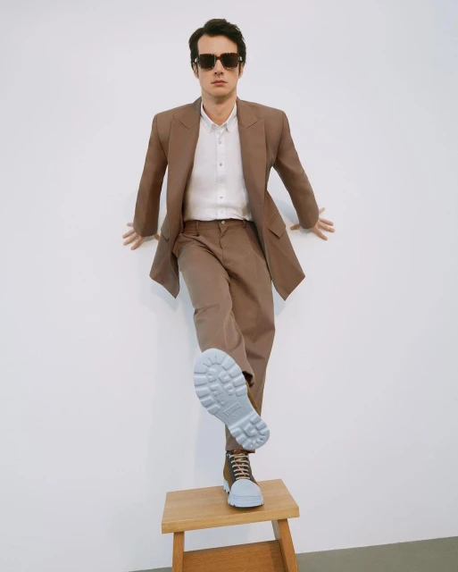 Stylish man in a brown suit posing on a wooden stool, showcasing a boot sole against a white backdrop in a fashion forward studio.
