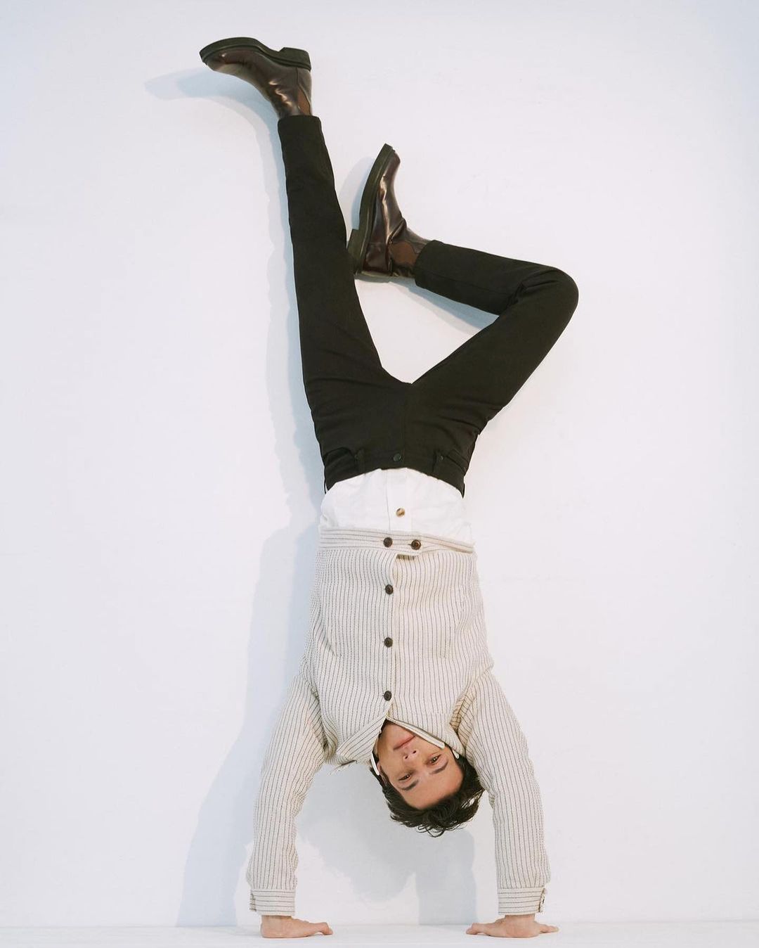 Adult male does a handstand against a white wall wearing black pants, brown leather boots, a white collared shirt, and a striped jacket.