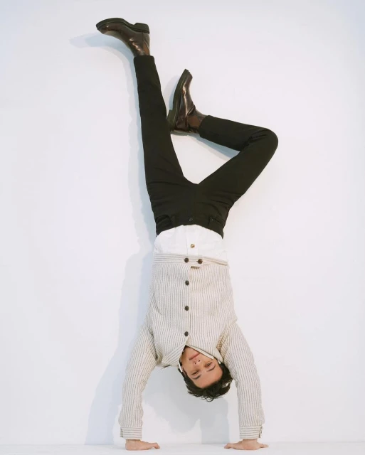 Adult male does a handstand against a white wall wearing black pants, brown leather boots, a white collared shirt, and a striped jacket.