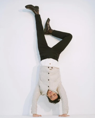 Adult male does a handstand against a white wall wearing black pants, brown leather boots, a white collared shirt, and a striped jacket.