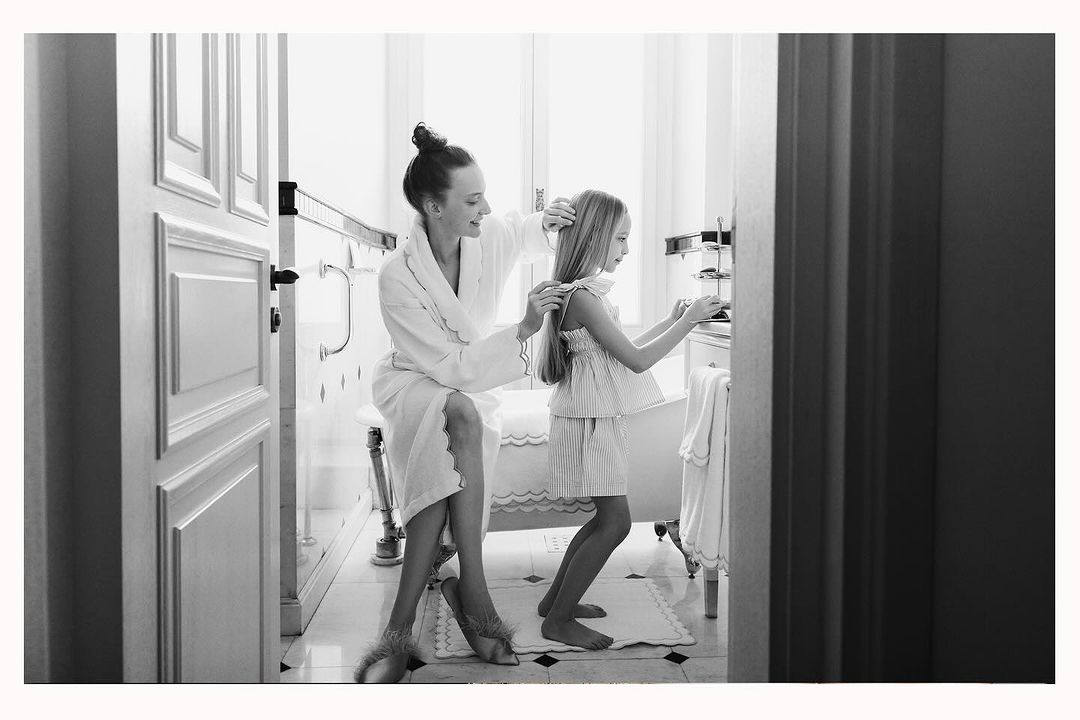 A black and white indoor shot of a woman in a robe seated on a bench, fixing the hair of a standing child near a bathroom vanity.