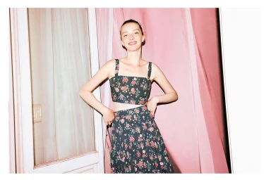 A smiling model in a floral two-piece set posing in front of pink drapes and a doorway, photographed by Memet Erol.