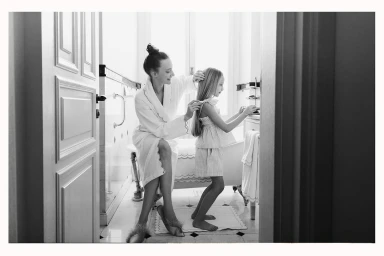 Woman in white bathrobe styling hair of a young girl in a bathroom captured in black and white by Memet Erol