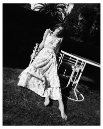 Monochrome portrait of a woman in a floral two-piece set sitting by a white table on grass outdoors.